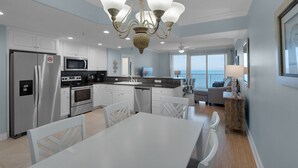 A modern kitchen with white cabinets, stainless steel appliances, and a large island. There is a dining area with white chairs and a table. The room has a large window with a view of the ocean. - 1906-19th Floor-PET FRIENDLY-Amazing Gulf View!-BEACH CHAIRS UMBRELLA INCL (Panama City Beach)