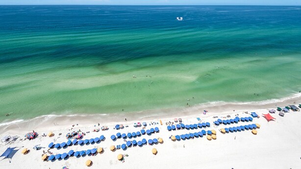 An aerial view of a beach with rows of blue and yellow sun loungers and umbrellas, clear turquoise water, and a few people in the distance. - 1906-19th Floor-PET FRIENDLY-Amazing Gulf View!-BEACH CHAIRS UMBRELLA INCL (Panama City Beach)