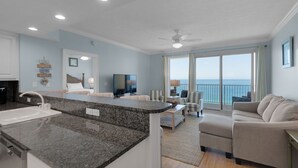 A modern living room with a kitchen area, a large flat-screen TV, and a balcony with a view of the ocean. - 1906-19th Floor-PET FRIENDLY-Amazing Gulf View!-BEACH CHAIRS UMBRELLA INCL (Panama City Beach)