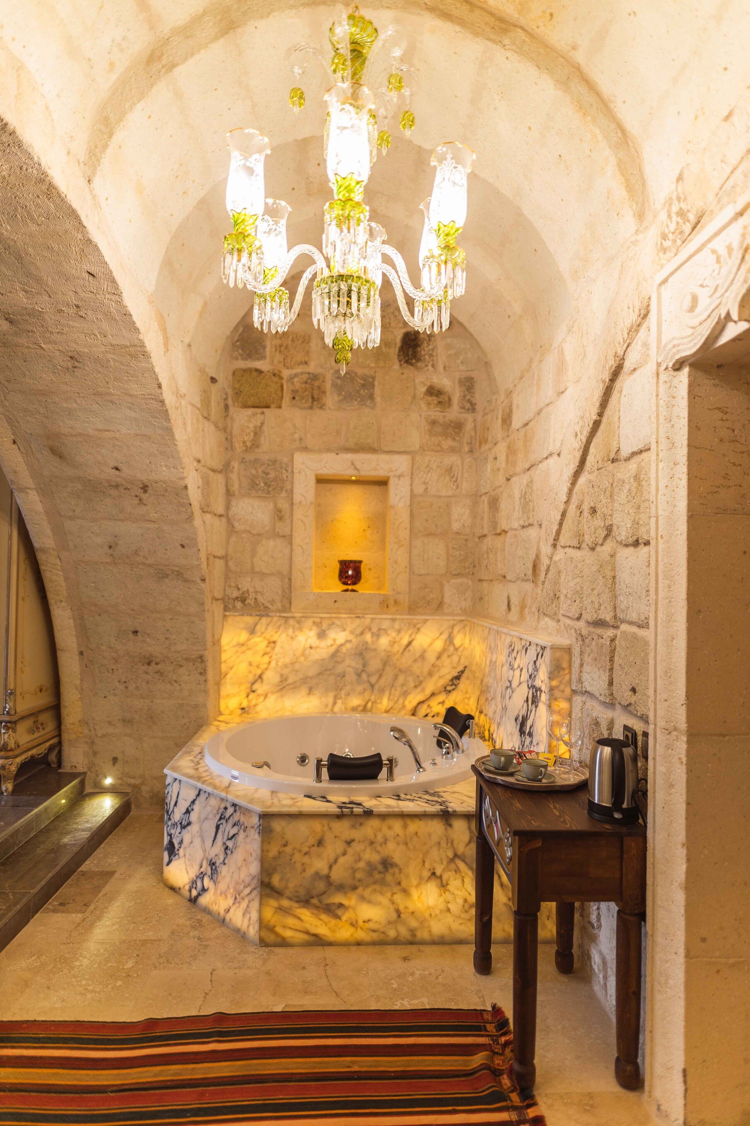 Un baño con paredes de piedra, una bañera de mármol, una mesita de madera con una tetera y un candelabro.