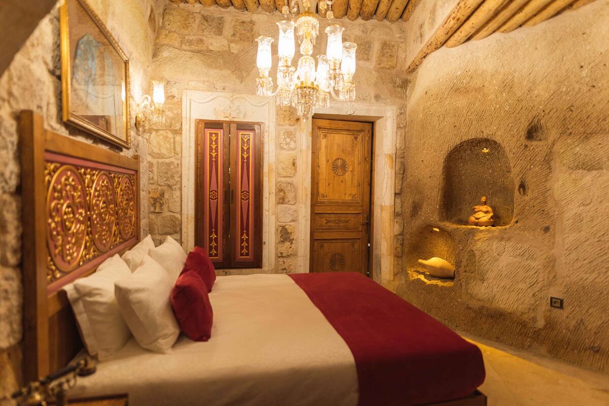 A room with a stone wall, a wooden door, a chandelier, and a bed with a red blanket.