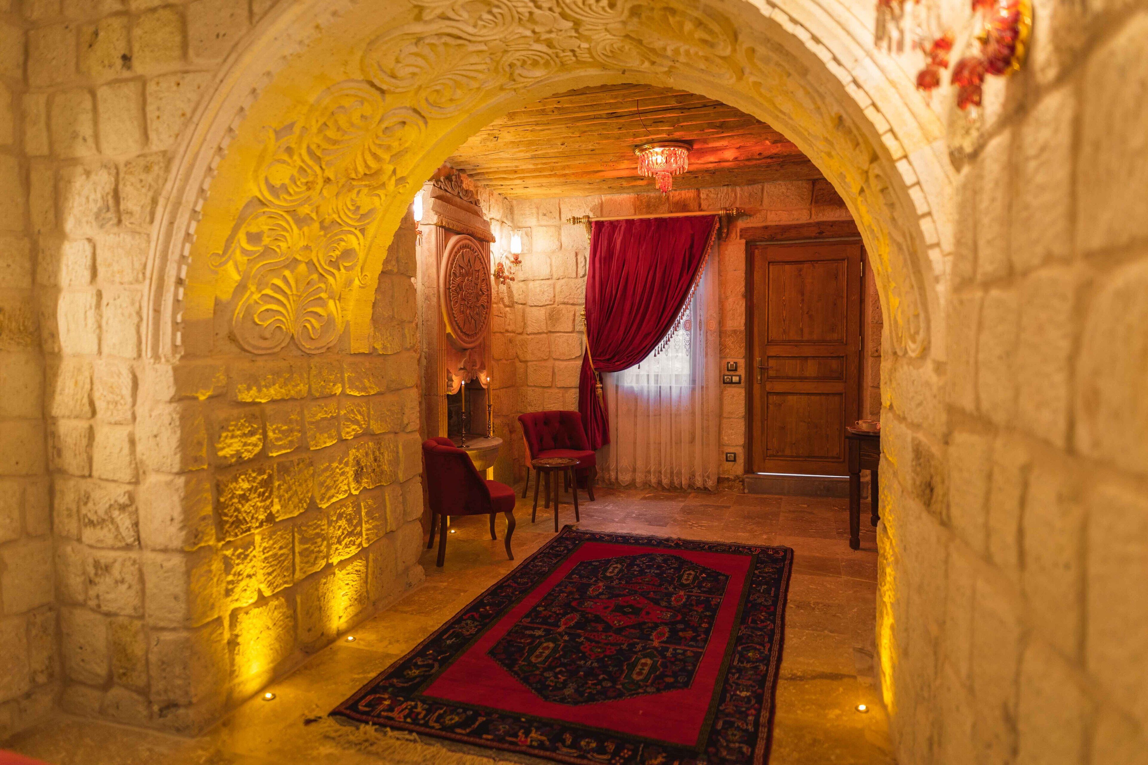 An ornate stone archway leading to a room with a red carpet, two chairs, and a wooden door.