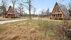Two wooden cabins with pitched roofs, surrounded by dry grass and trees.