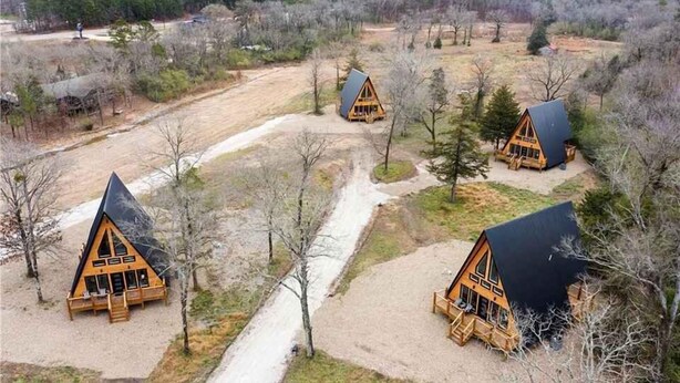 Three wooden cabins with pitched roofs, surrounded by trees and a dirt path.