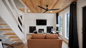 A modern living room with a wooden ceiling, a flat-screen TV mounted on the wall, a sofa, and a staircase with white railings. - “Elk” High LUXE 1 Bedroom A-Frame, The Alps of Hochatown (Broken Bow)