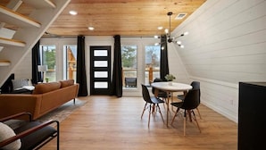 A modern loft-style room with a wooden ceiling, a dining area with a round table and chairs, a sofa, and a staircase. - “Elk” High LUXE 1 Bedroom A-Frame, The Alps of Hochatown (Broken Bow)