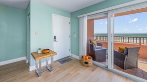 Ein Zimmer mit Holzboden, einer weißen Tür, einem kleinen Tisch mit einer Pflanze, einem Korbstuhl und Blick auf das Meer durch eine Schiebeglastür.