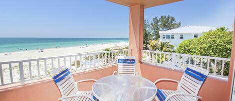 A balcony with a table and chairs, overlooking a beach with a clear view of the ocean and a building in the background.