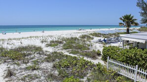 Ein Strand mit weißem Sand, klarem blauen Wasser und einigen Sonnenschirmen. Liegestühle und eine Palme sind ebenfalls vorhanden.