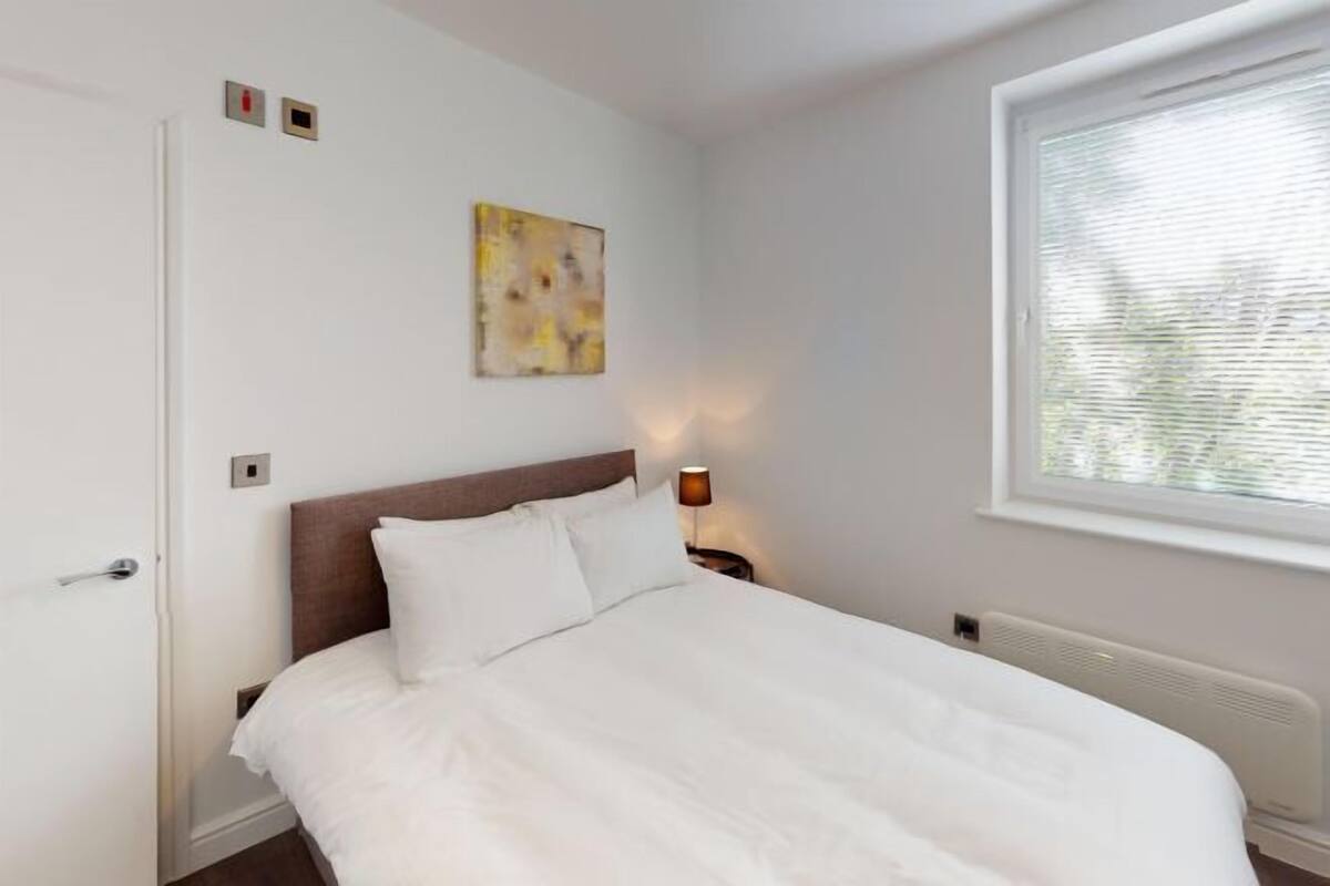 A bedroom with a bed, a nightstand, a wall-mounted lamp, a window with blinds, and a door.