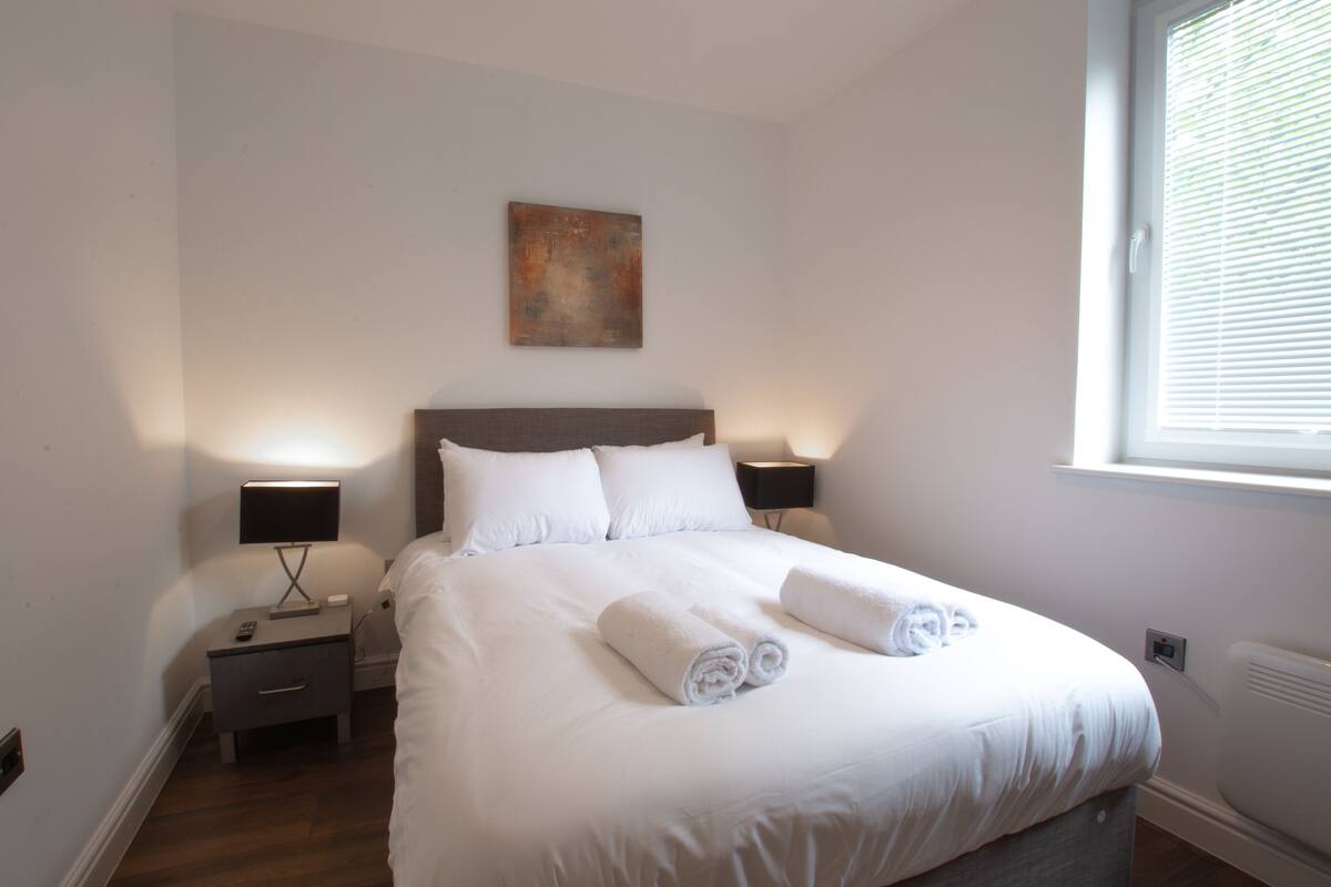A neatly made bed with white linens and towels, two bedside tables with lamps, a wall-mounted artwork, and a window with blinds.