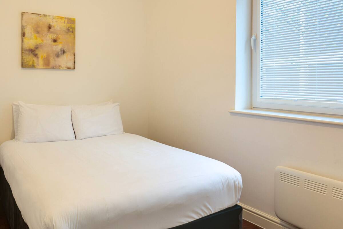 A bedroom with a bed, a nightstand, a lamp, a window with blinds, and a painting on the wall.