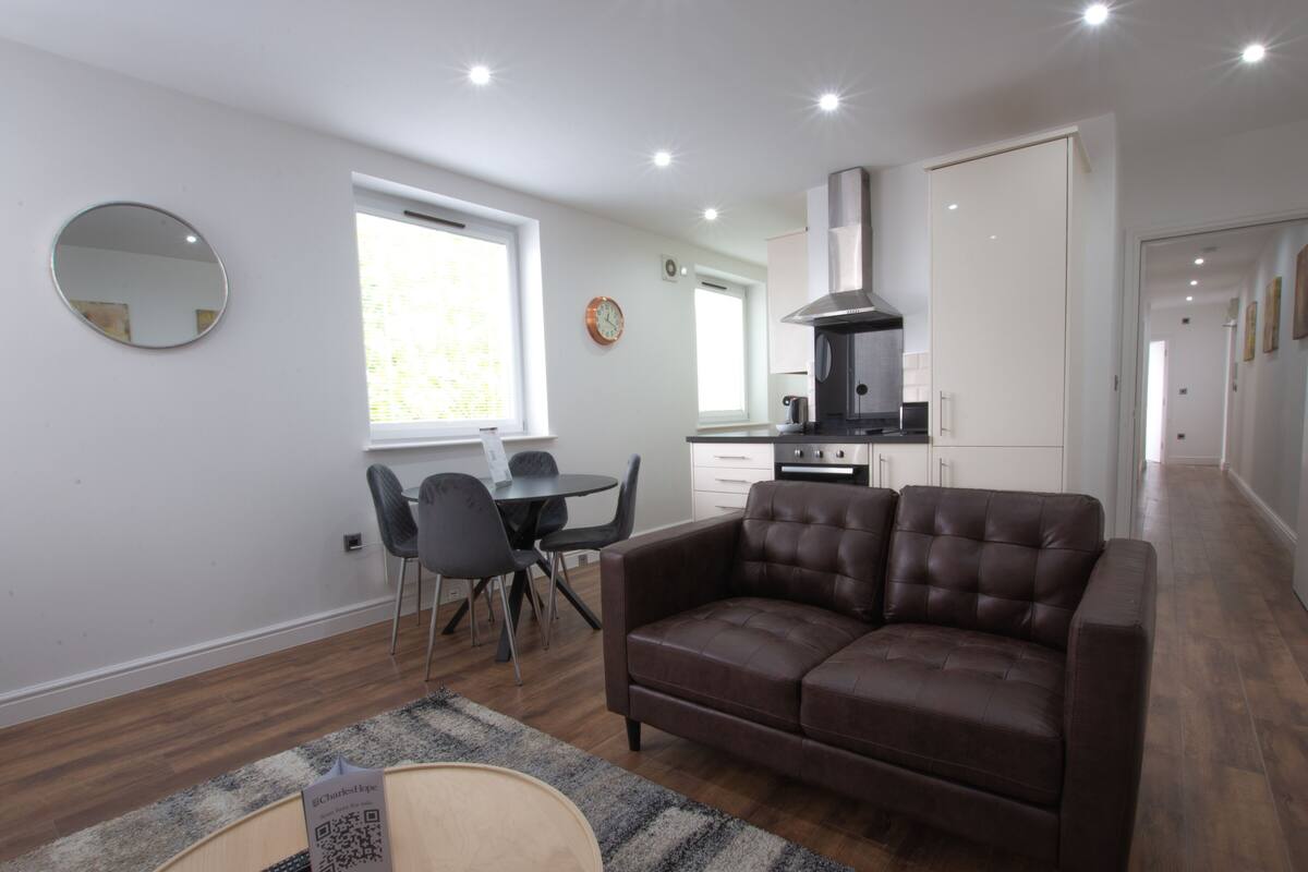 A modern living room with a brown leather sofa, a dining table with chairs, a kitchen area with a stove and oven, and a large window providing natural light.
