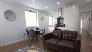 A modern living room with a brown leather sofa, a dining table with chairs, a kitchen area with a stove and oven, and a large window providing natural light. - Charles Hope Apartments Swindon (Swindon)