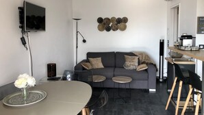 Ett modernt vardagsrum med en grå soffa, ett matbord med stolar, en tv som är monterad på väggen och en kaffemaskin på en bänkskiva.