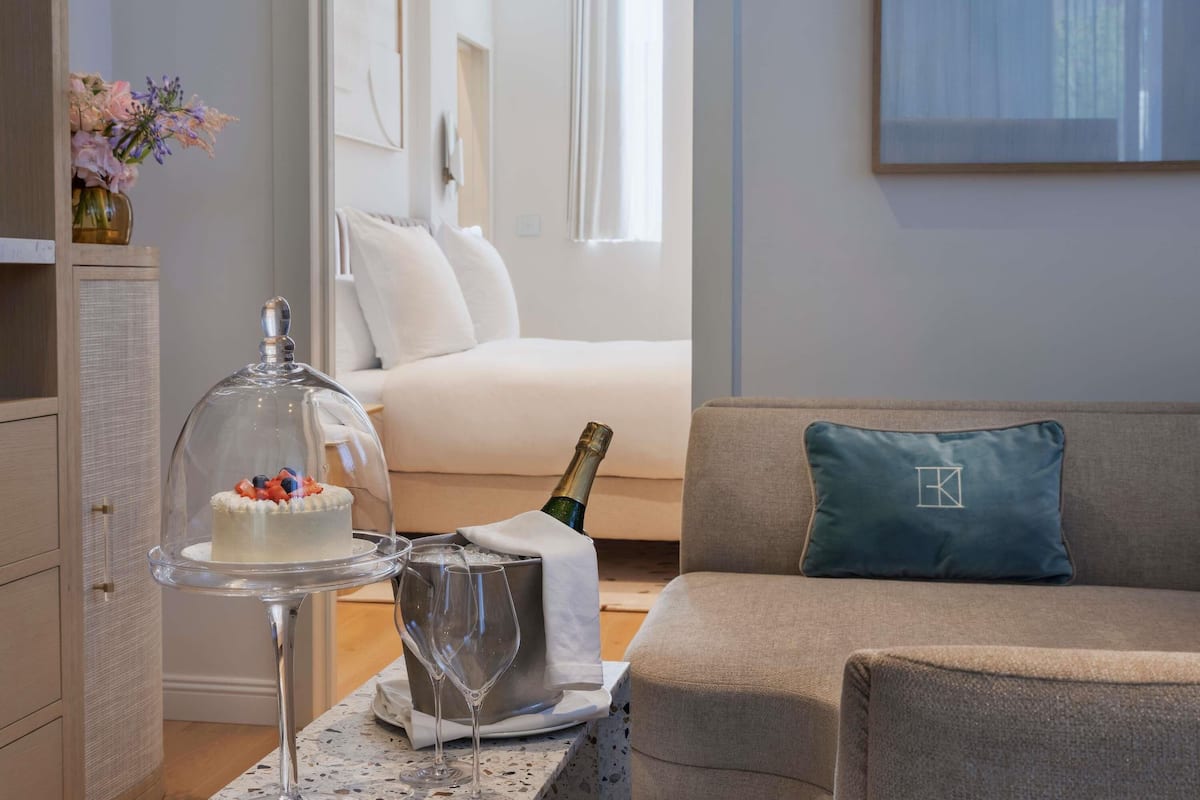 Una stanza con un letto, una poltrona, un vassoio per dolci in vetro, una bottiglia di champagne e un vaso decorativo.