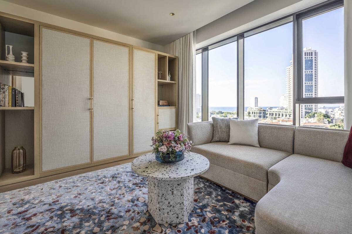 Una sala de estar moderna con un sofá, una mesa redonda de centro con un arreglo floral, una estantería y un ventanal con vista a la ciudad.