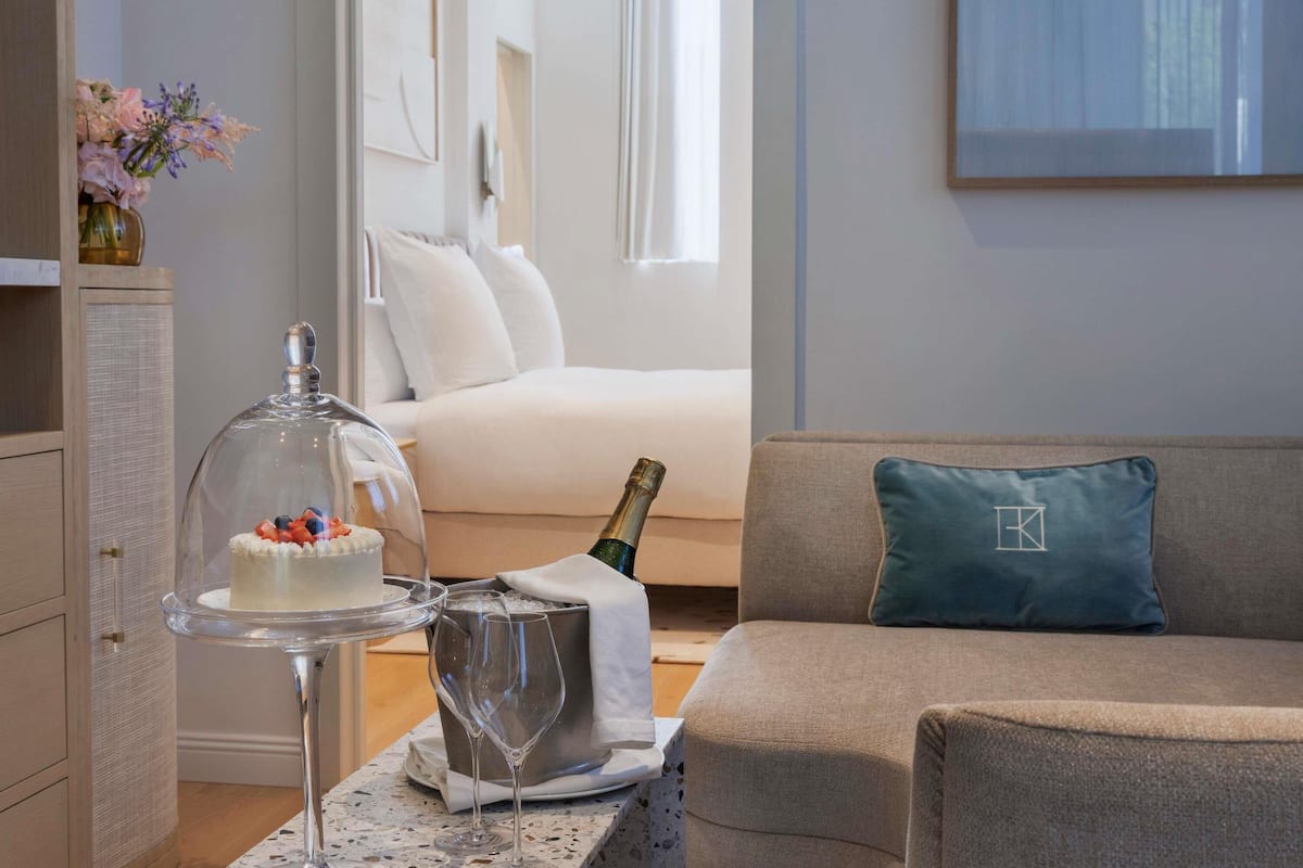 Una habitación con cama, un sofá, un pastelero de cristal con un pastel, una botella de champán y un jarrón decorativo.