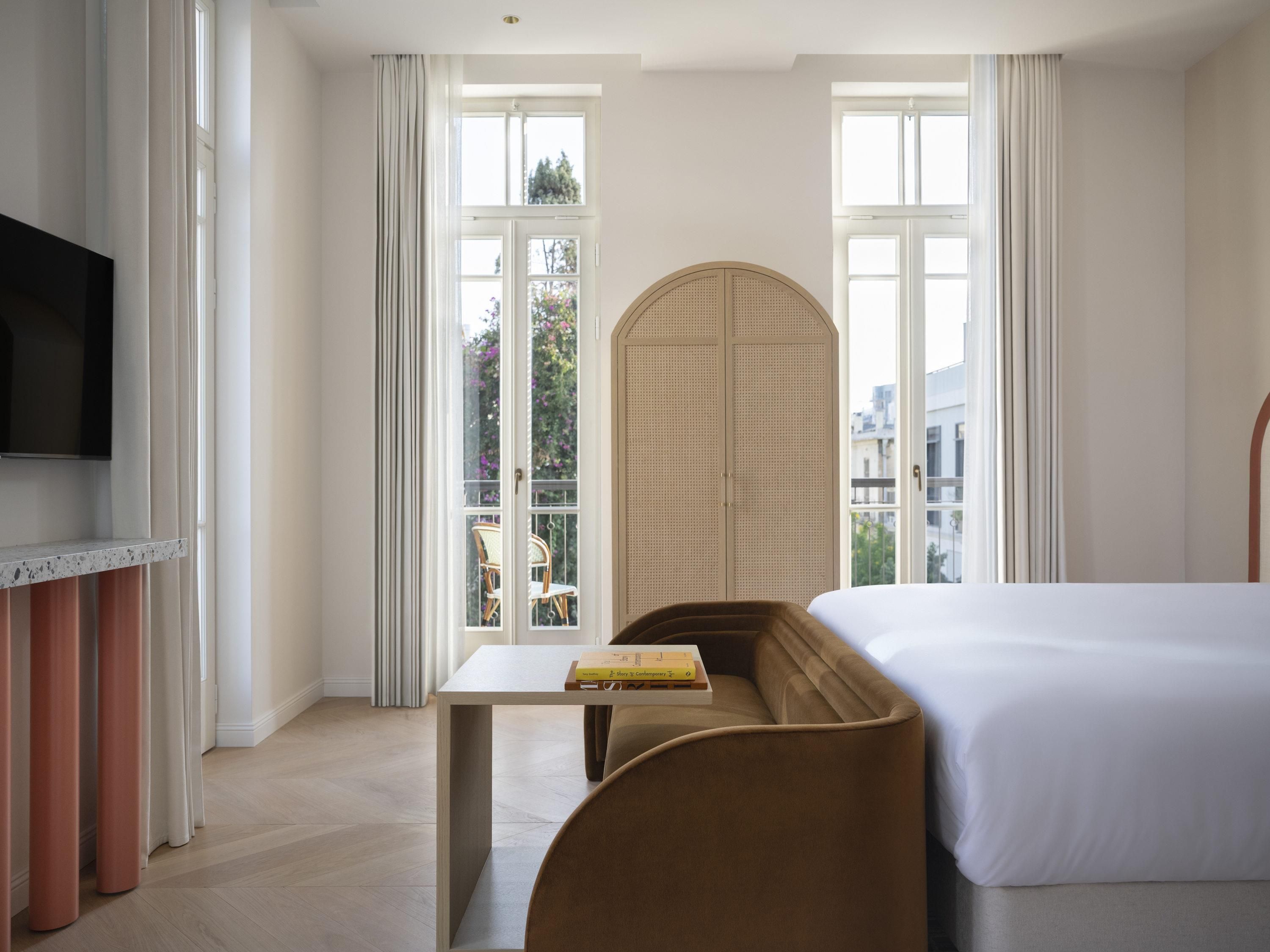 Habitación de hotel con cama, televisión, una mesa con libros y vistas al exterior a través de amplias ventanas.