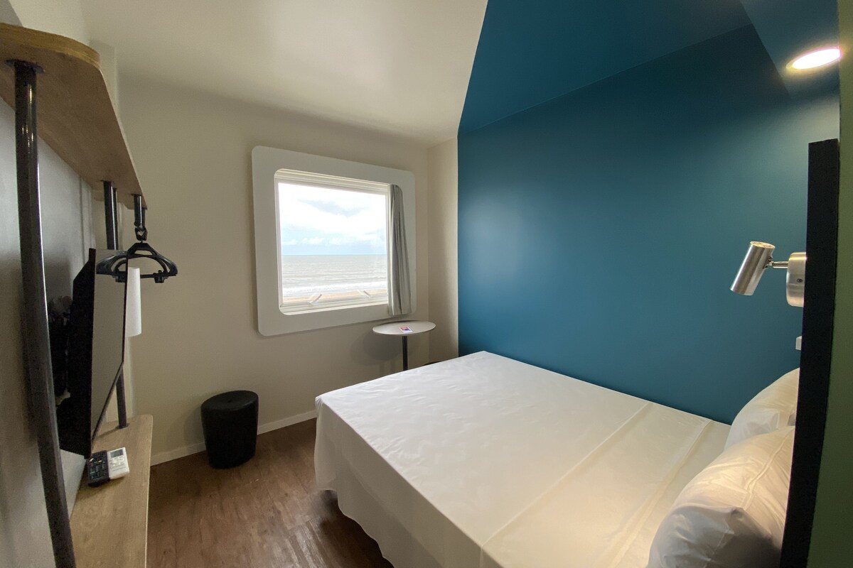 A bedroom with a bed, a window with a view of the beach, a small table, and a trash bin.