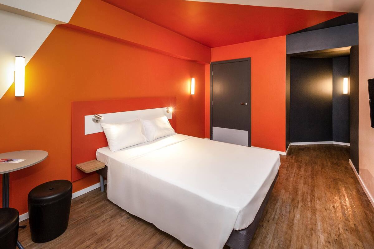 A hotel room with a bed, a desk, and a chair. There is a door and a light fixture on the wall.