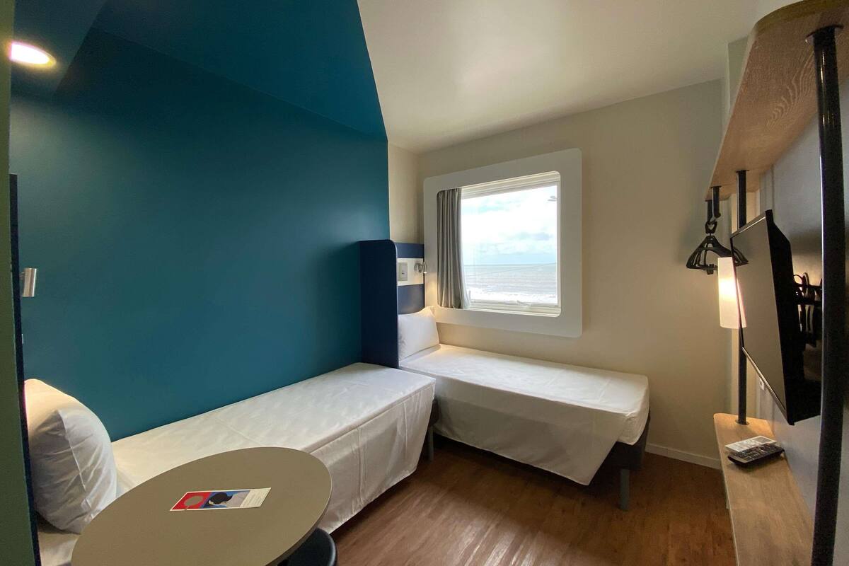 A compact room with two single beds, a small table, a window with a view of the sea, and a wall-mounted TV.