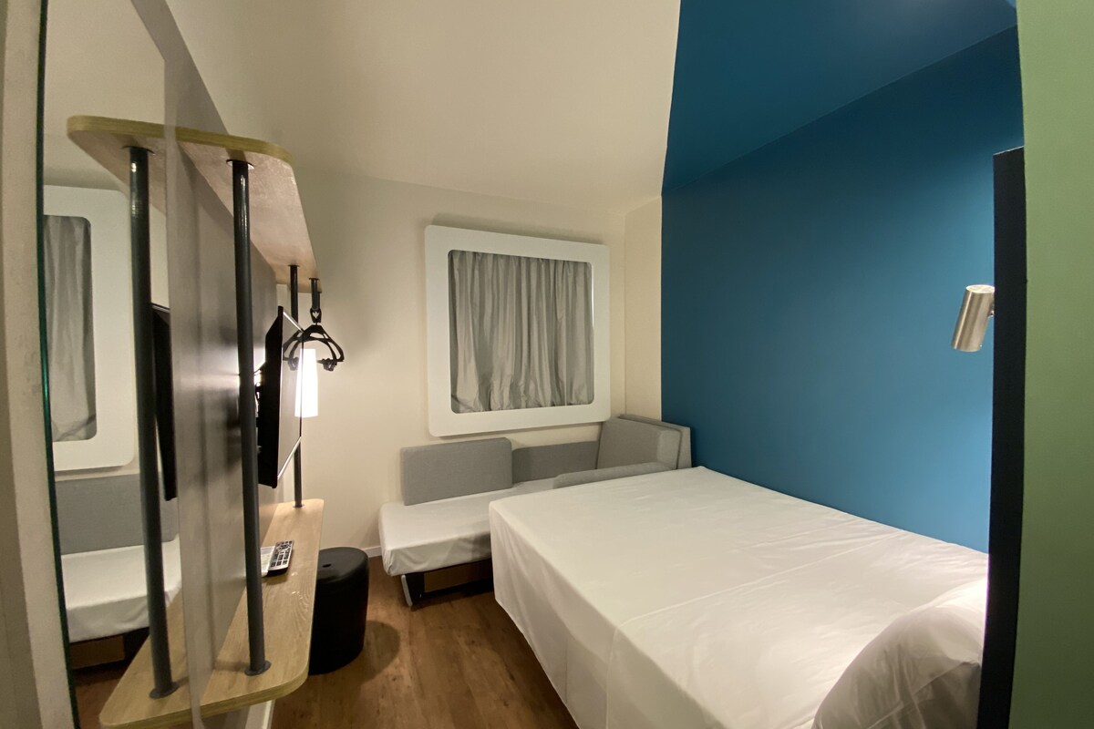 A hotel room with a bed, a small seating area, a window with curtains, a mirror, and a wall-mounted light fixture.
