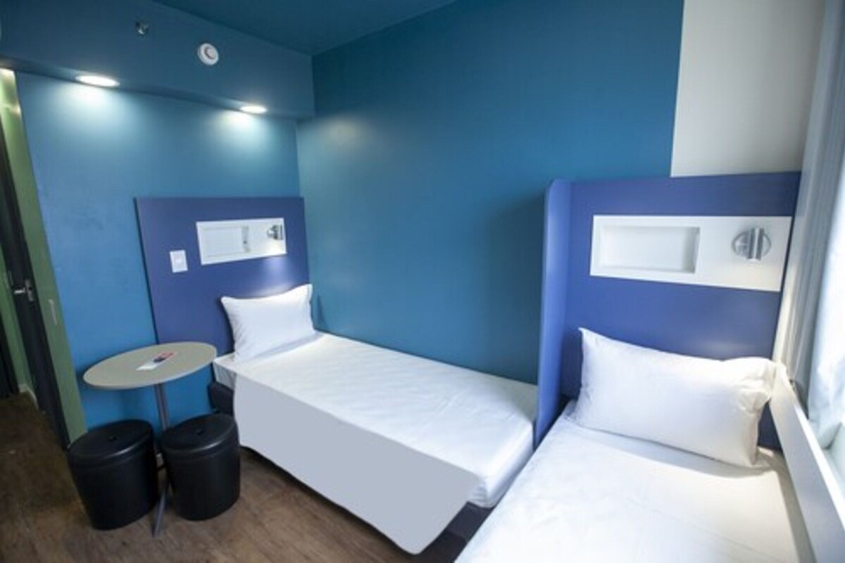 A hotel room with two single beds, a small round table, and two trash bins.
