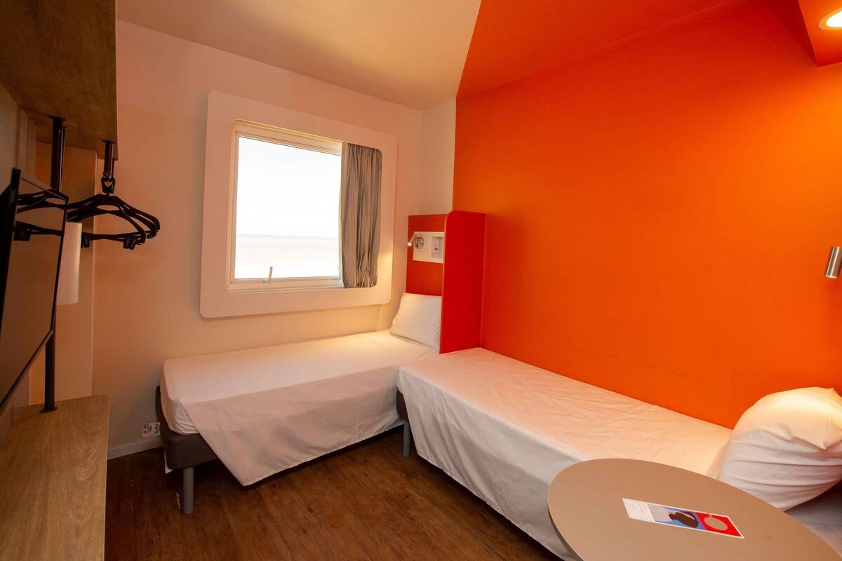 A room with two beds, a small table, a window with curtains, and a coat rack.