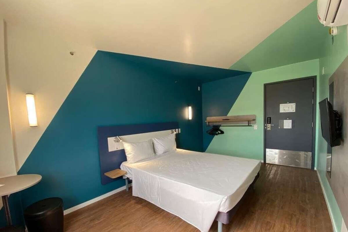 A single bed in a room with a sloped ceiling, a small table, a trash bin, and a door with a mirror.