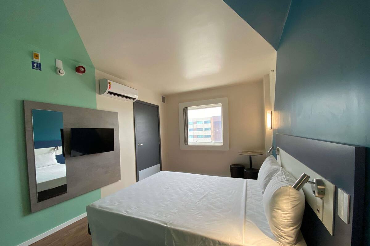 A hotel room with a bed, a mounted TV, a window with a view of buildings, and a wall-mounted air conditioning unit.