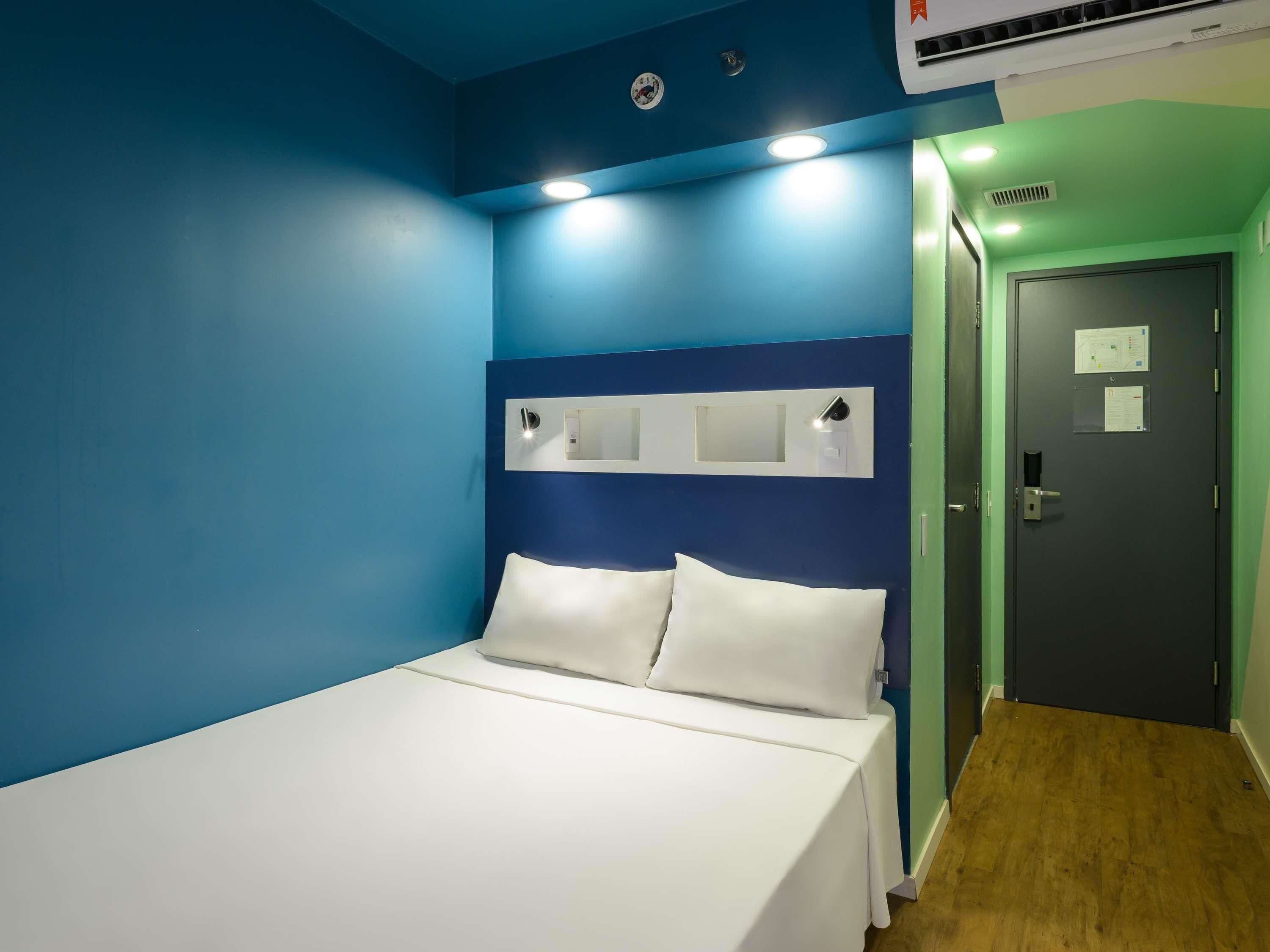Habitación de hotel con cama, mesita de noche, luz empotrada en la pared y puerta con cerradura.