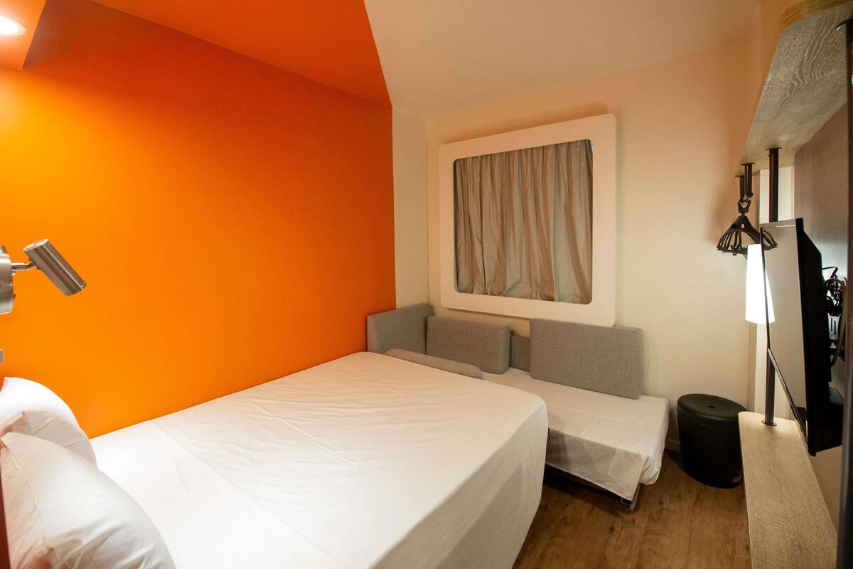 Ein Zimmer mit oranger Wand, einem Bett, einem kleinen Sofa, einem Schreibtisch mit Lampe und einem fenster mit Vorhängen.