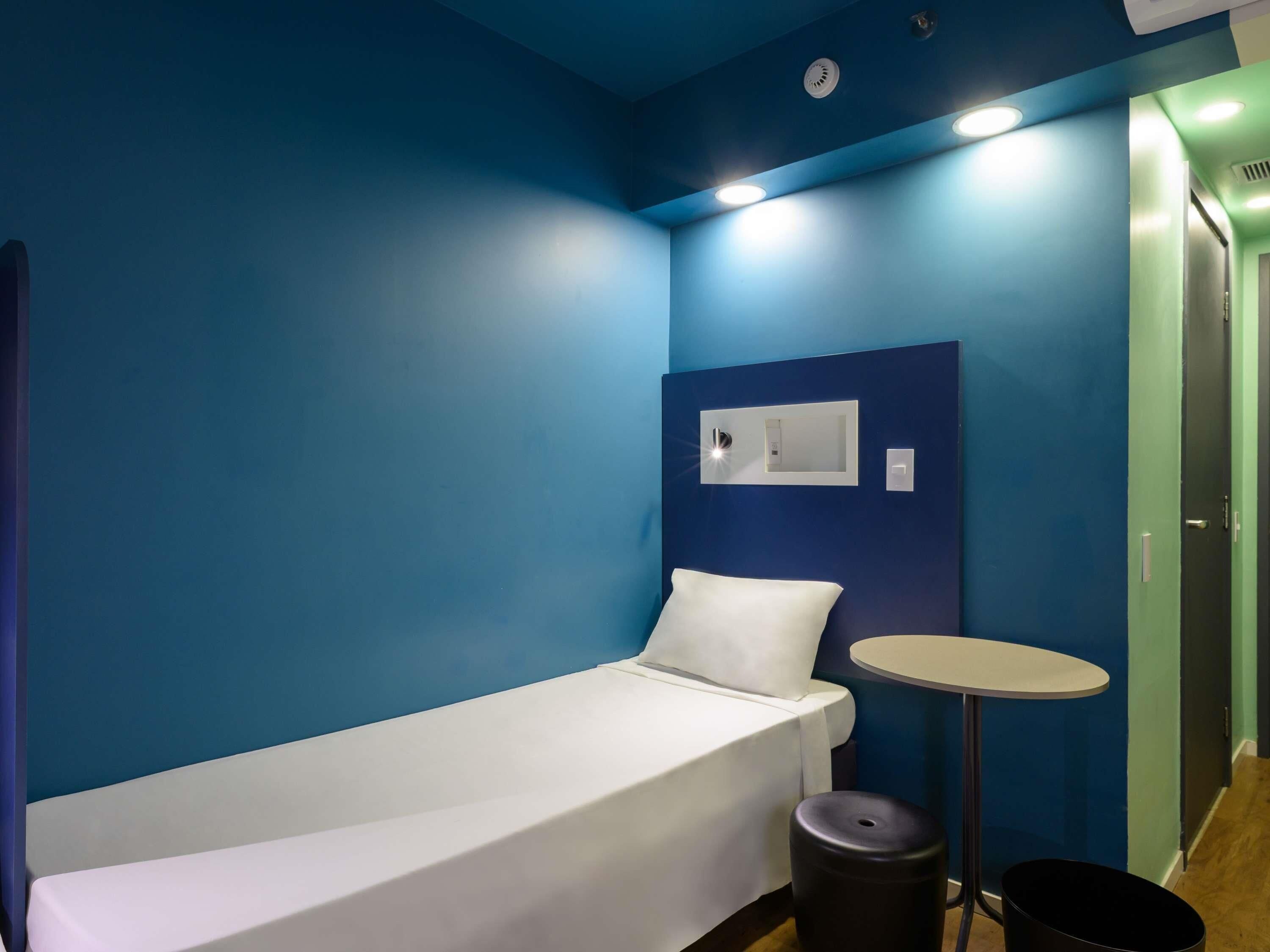 A single bed with a white mattress, a small round table, a black trash bin, and a wall-mounted light switch.