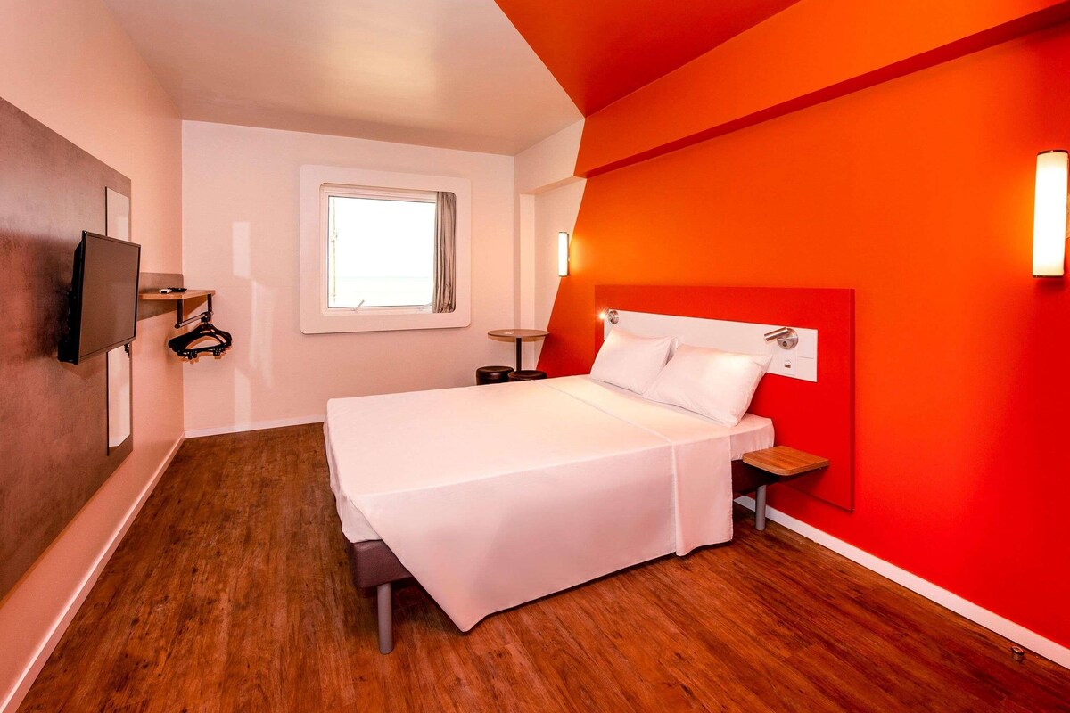 Une chambre d’hôtel avec un lit, un téléviseur fixé au mur, une fenêtre avec des rideaux et un mur d’accent de couleur rouge.