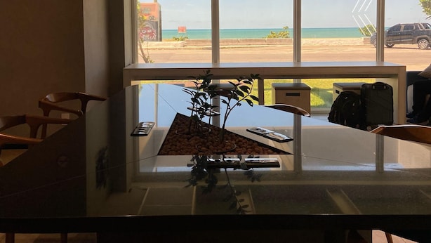 A hotel lobby with a large window offering a view of the beach. There is a reception desk with a plant on it, and a few chairs around the desk. - ibis budget Recife Jaboatão (Jaboatão dos Guararapes)