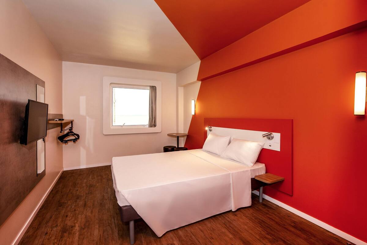 A hotel room with a bed, a TV mounted on the wall, a window, and a red accent wall.