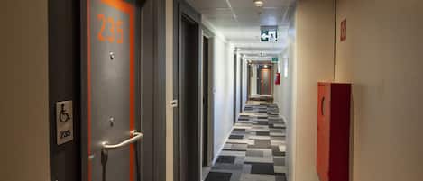 Pasillo de hotel con suelo a cuadros, un detector de incendios y señal de accesibilidad para sillas de ruedas.