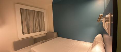 Una habitación de hotel compacta con una cama individual, un pequeño banco, una ventana con cortinas y una lámpara fijada a la pared.
