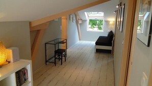 Ein Loft-Zimmer mit schrägem Dach, einem kleinen Tisch mit Stuhl, einer Couch und einem Fenster mit Blick auf Grünflächen.