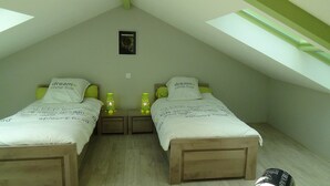Ein kleines, gemütliches Schlafzimmer im Dachgeschoss mit zwei Einzelbetten, jedes mit einem Kopfkissen mit dem Aufdruck „dreams come true“. Es gibt Nachttische mit grünen Lampen und ein kleines gerahmtes Bild an der Wand.