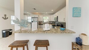 A modern kitchen with white cabinets, granite countertops, and a central island with stools. - Wave n Sea - Close to the Beach with Pool - Anna Maria Island - By Island Real E (Anna Maria)