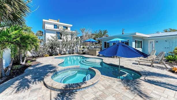 A swimming pool with a tiled edge, surrounded by a patio area with lounge chairs and umbrellas. - Wave n Sea - Close to the Beach with Pool - Anna Maria Island - By Island Real E (Anna Maria)