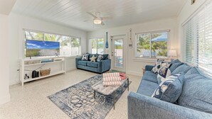 A spacious living room with a blue sofa, a white entertainment unit, and large windows. - Wave n Sea - Close to the Beach with Pool - Anna Maria Island - By Island Real E (Anna Maria)