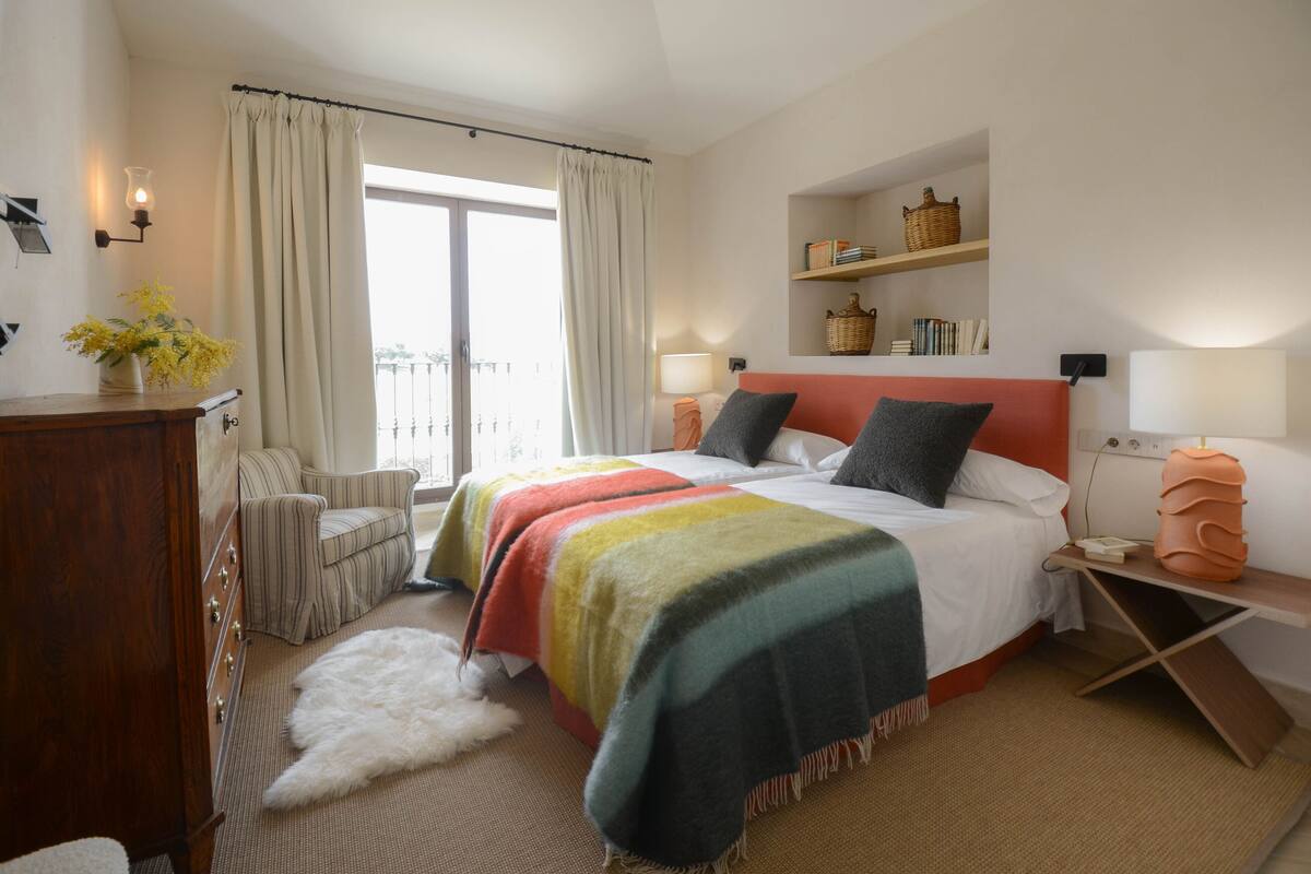 A hotel room with a bed, a chair, a dresser, a lamp, a shelf with books and a basket, and a view of the outdoors through a window.