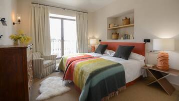 Habitación de hotel con cama, una silla, un escritorio, una lámpara, una estantería con libros y vistas al exterior.