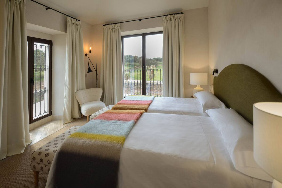 A hotel room with two beds, a chair, a lamp, and a view of the outdoors.