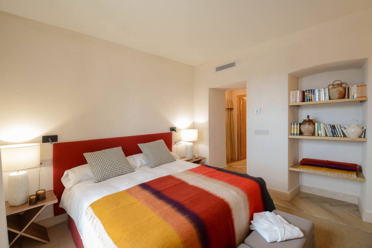 A hotel room with a bed, bedside lamps, a bookshelf, and a door leading to another room.