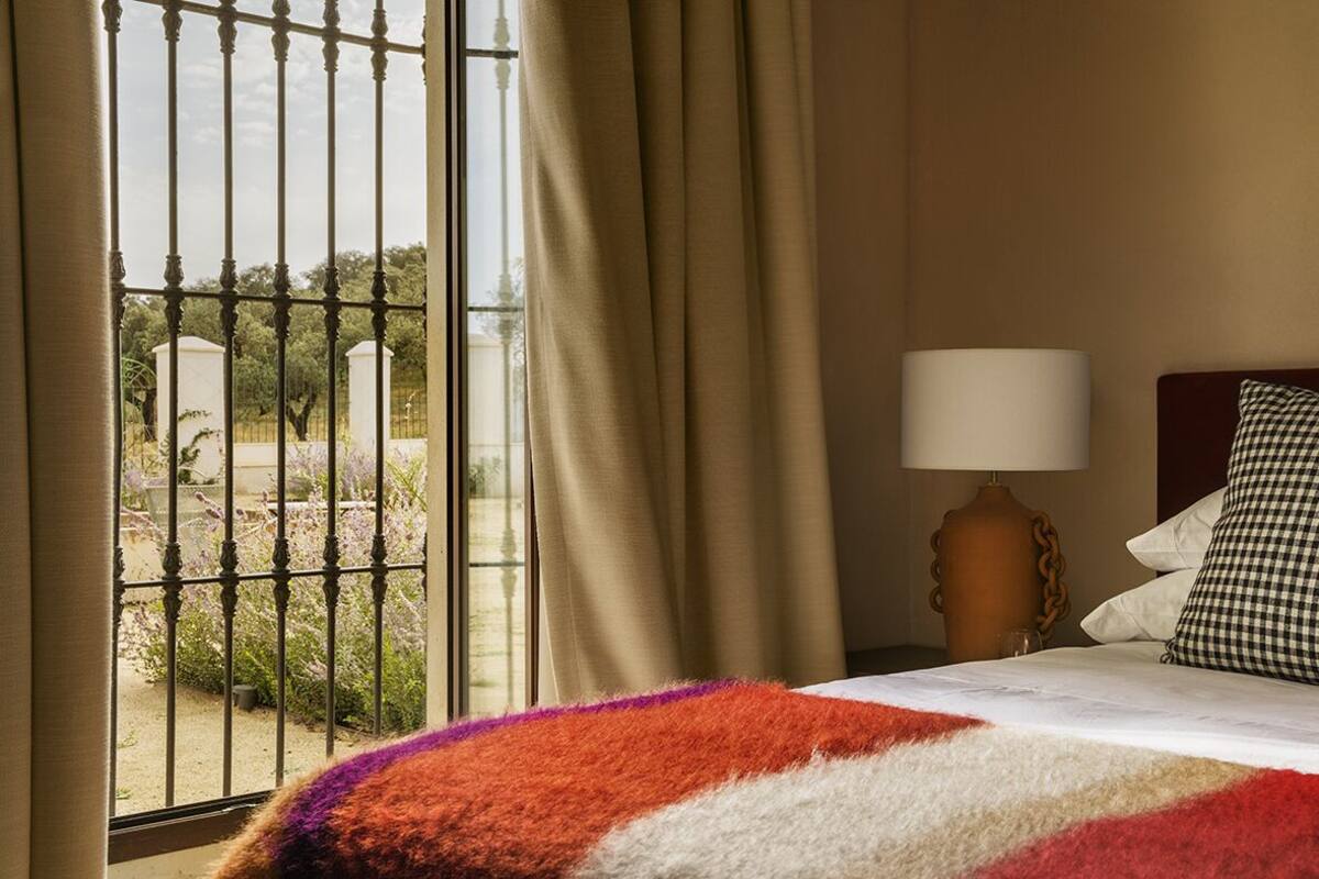 A hotel room with a bed, a lamp, and a view through a window with a metal gate.