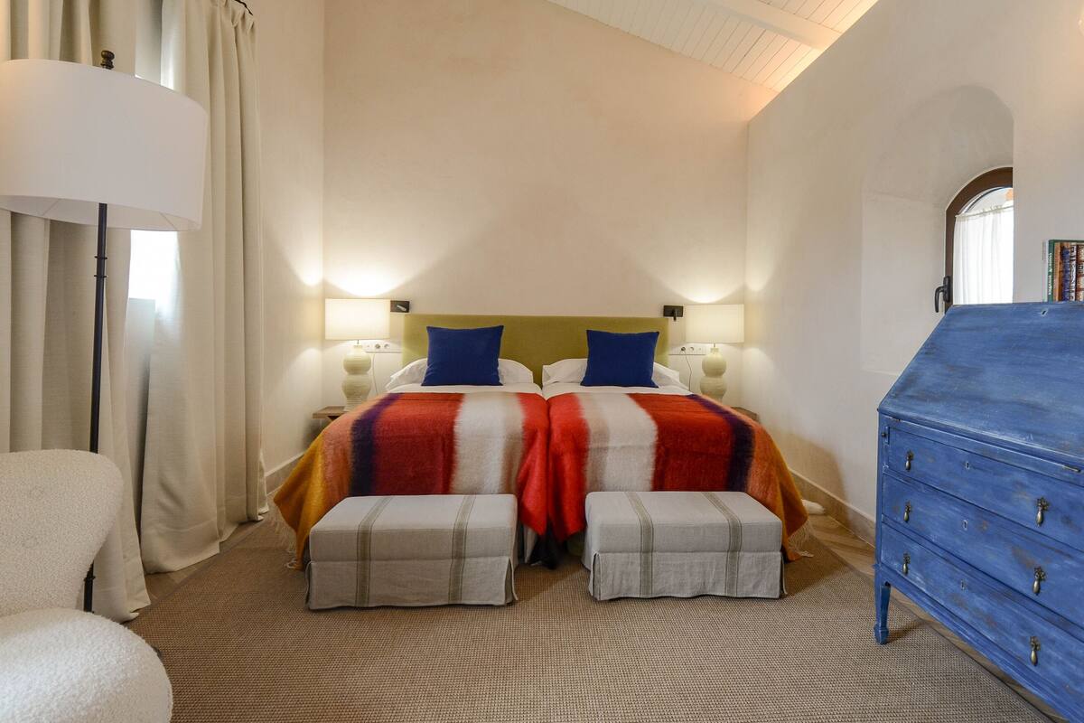 A double bed with a striped bedspread, two bedside tables with lamps, a blue dresser, and a large window with curtains.